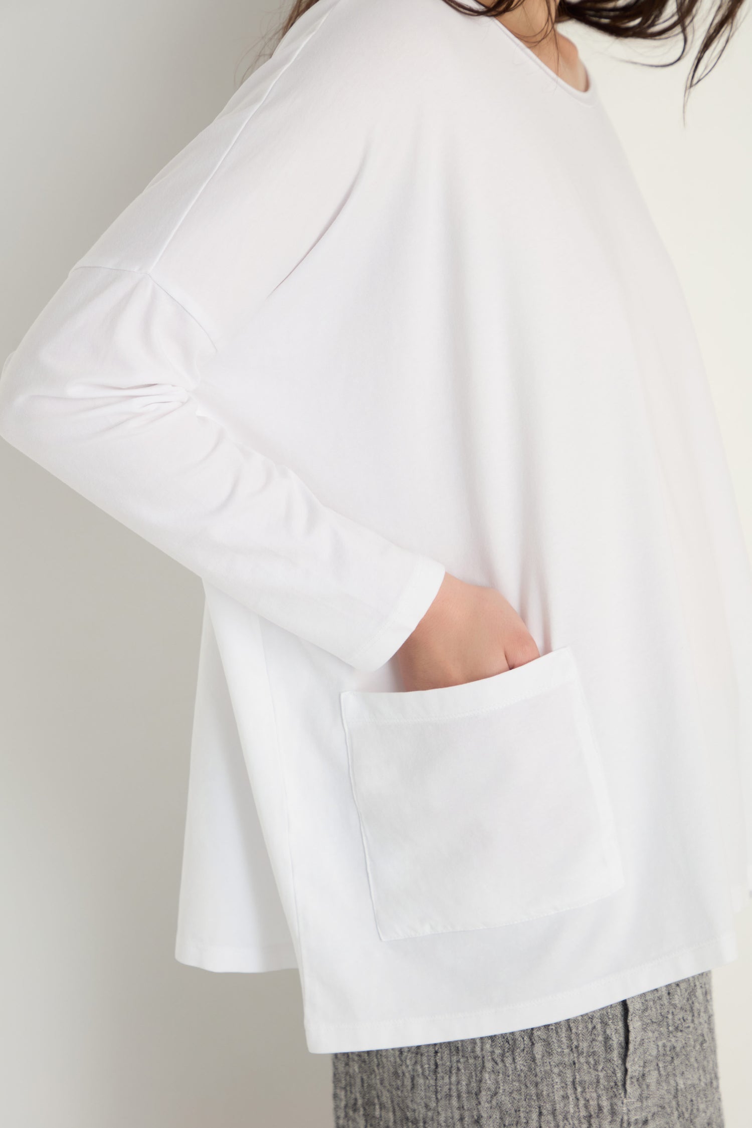 A person wearing a Cotton Jersey Boxy Pocket Top—a loose-fitting white long-sleeve with a front pocket—paired with gray textured pants.