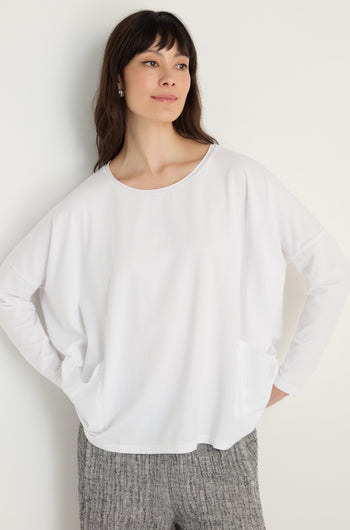 A woman stands against a white wall wearing the Cotton Jersey Boxy Pocket Top with a loose fit and front pockets, paired with gray textured pants.