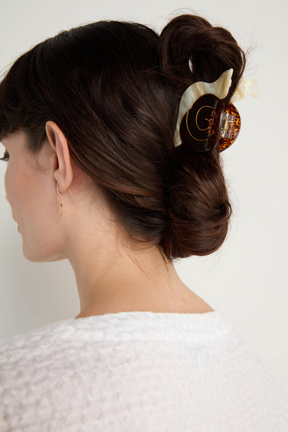 A woman with dark hair in a low bun, secured with a hand-painted Snail Hair Clip made from eco-friendly cellulose acetate, wears a white textured top and small hoop earring as she faces away from the camera.