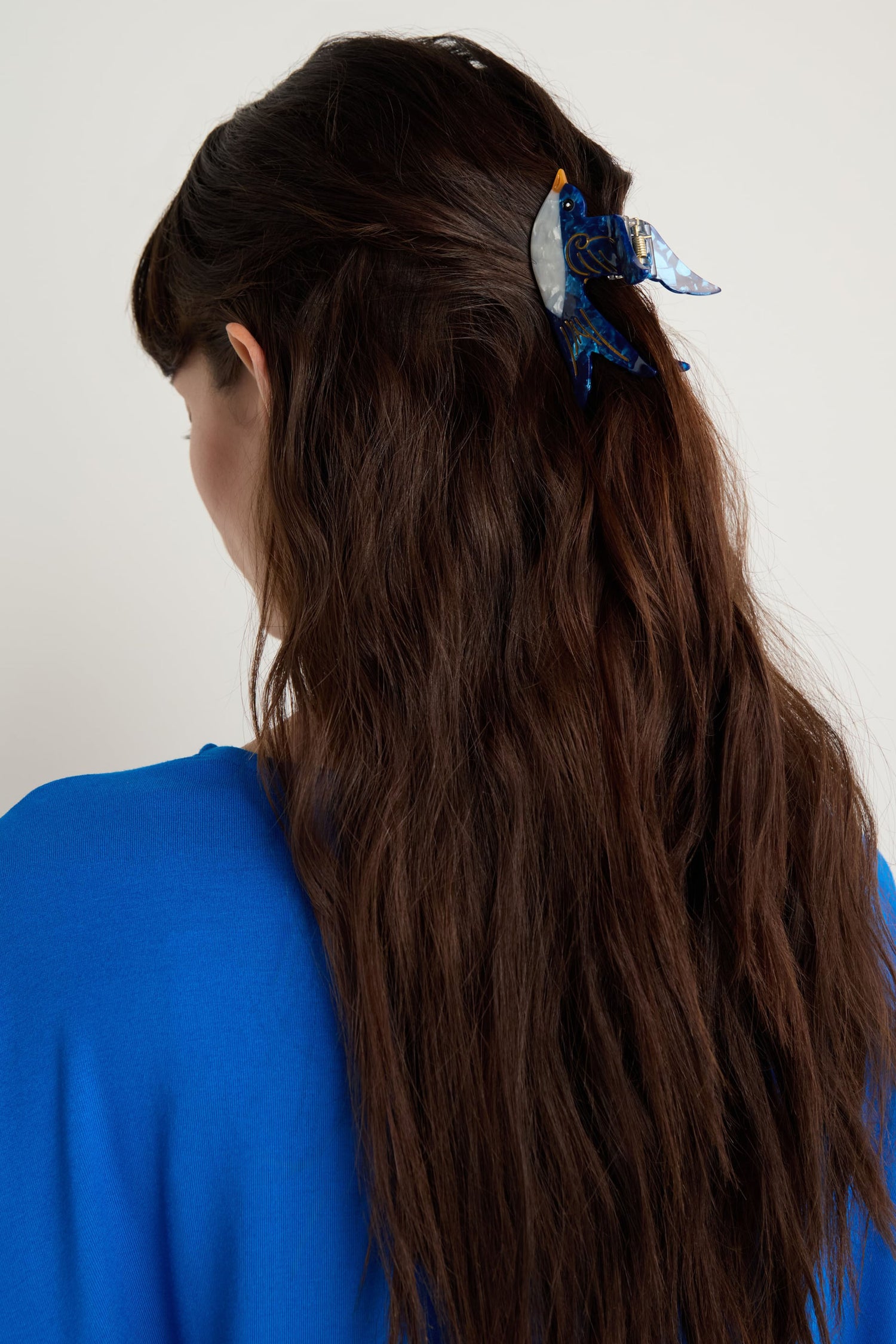 A woman with long, wavy brown hair wears a blue top and a Hand-Painted Swallow Hair Clip in blue and silver.
