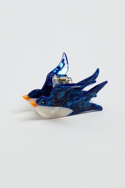 The Hand-Painted Swallow Hair Clip is an eco-friendly blue and white bird-shaped accessory with an orange beak and gold detailing, shown on a plain light background.