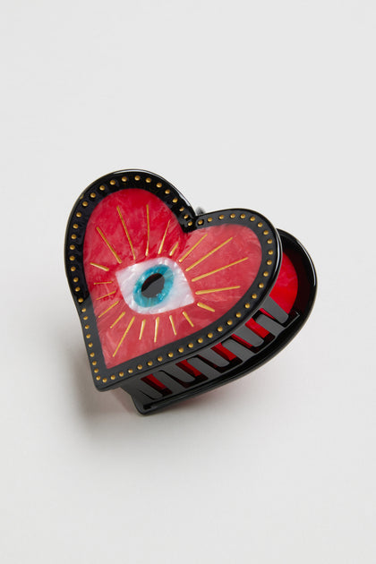 The Hand Painted Heart Hair Clip is crafted from biodegradable cellulose acetate, with a red hand-painted heart, gold rivets, and a blue eye with radiant gold rays at the center.