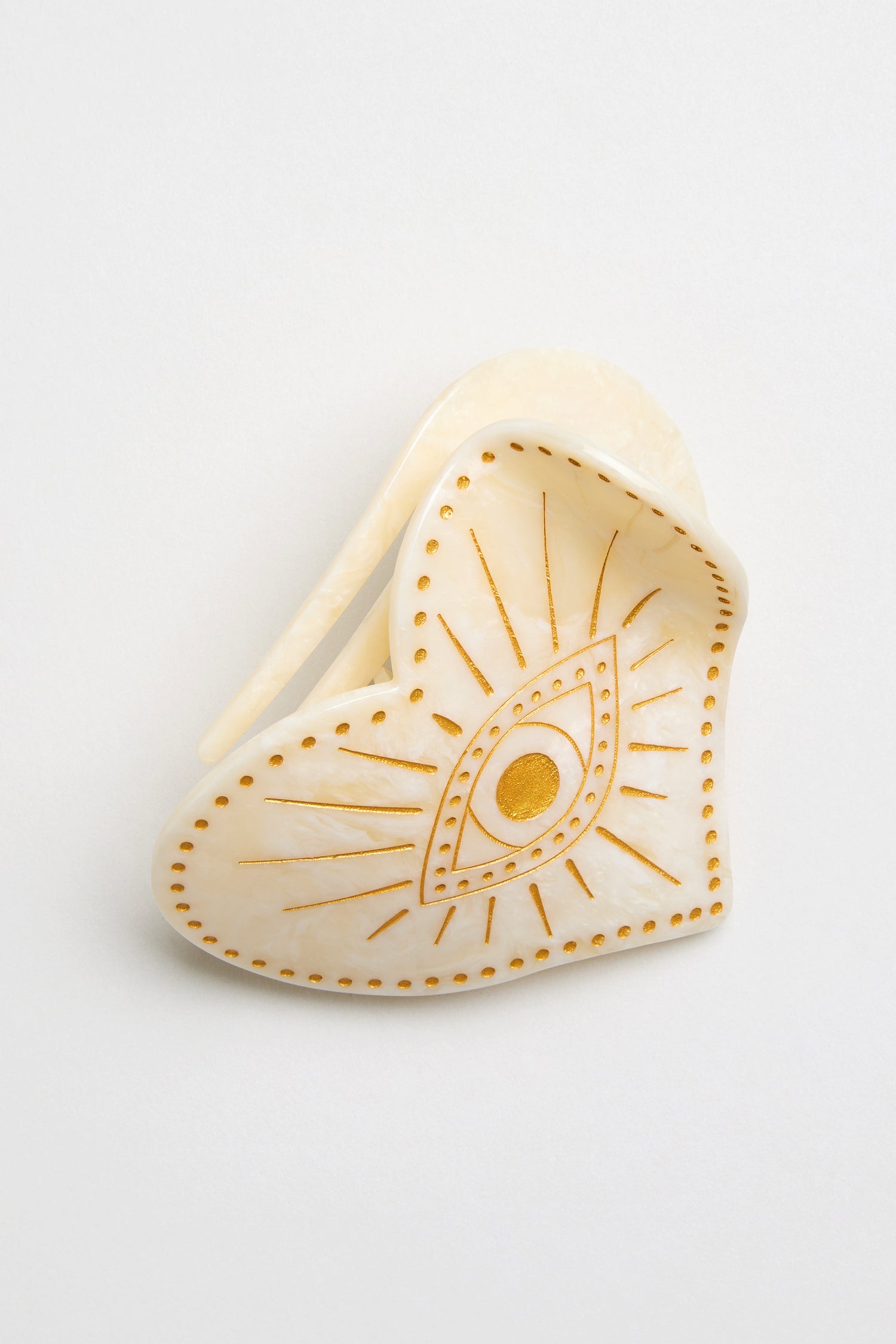 The Hand Painted Celestial Heart Hair Clip is crafted from cream biodegradable cellulose acetate, featuring a stylized gold eye with radiating lines and dotted accents.