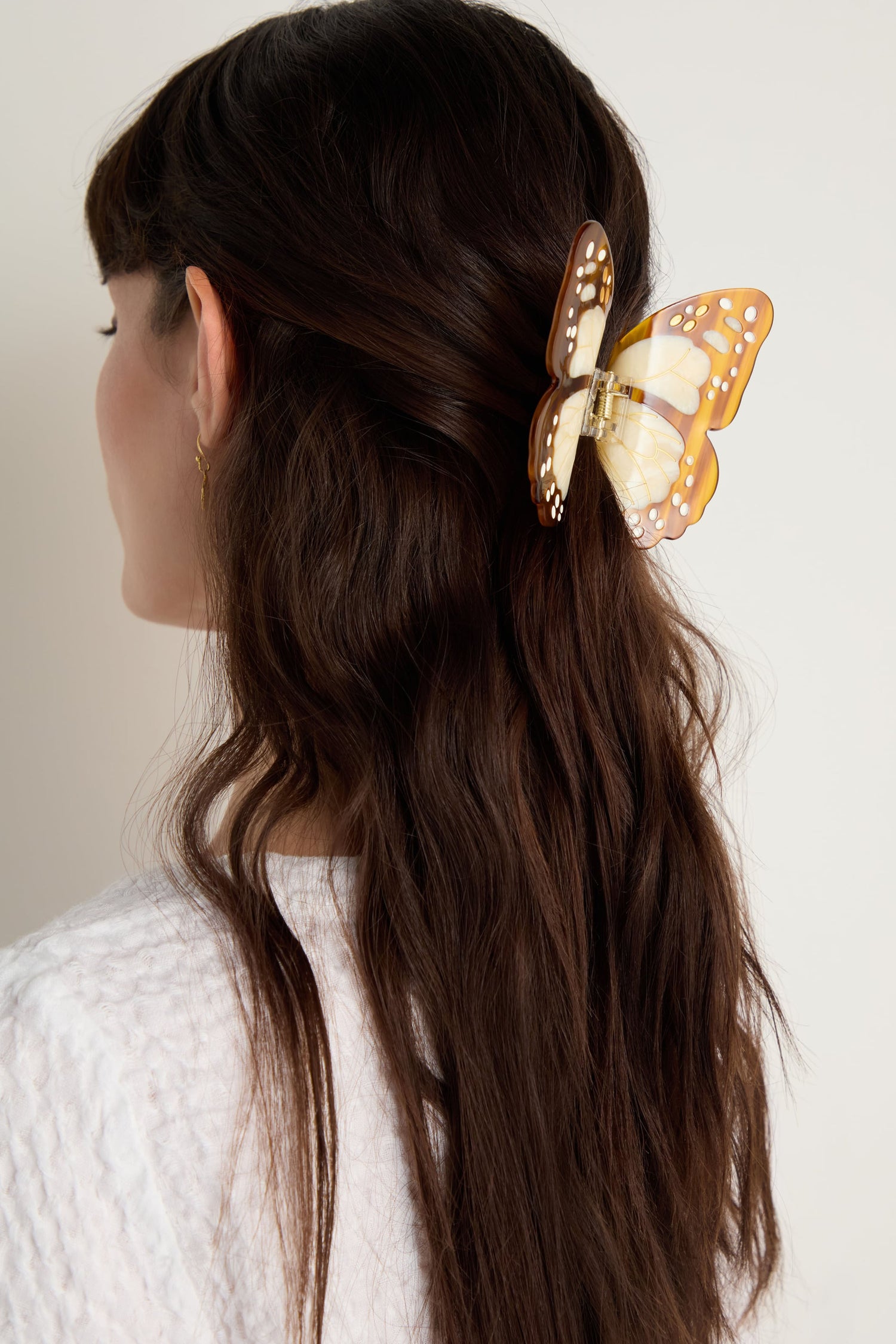 A woman with long brown hair wears a white textured top, styled with the Hand-painted Monarch Butterfly Hair Clip—an eco-friendly accessory that adds a unique touch to the back of her head.