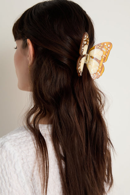 A woman with long brown hair wears a white textured top, styled with the Hand-painted Monarch Butterfly Hair Clip—an eco-friendly accessory that adds a unique touch to the back of her head.