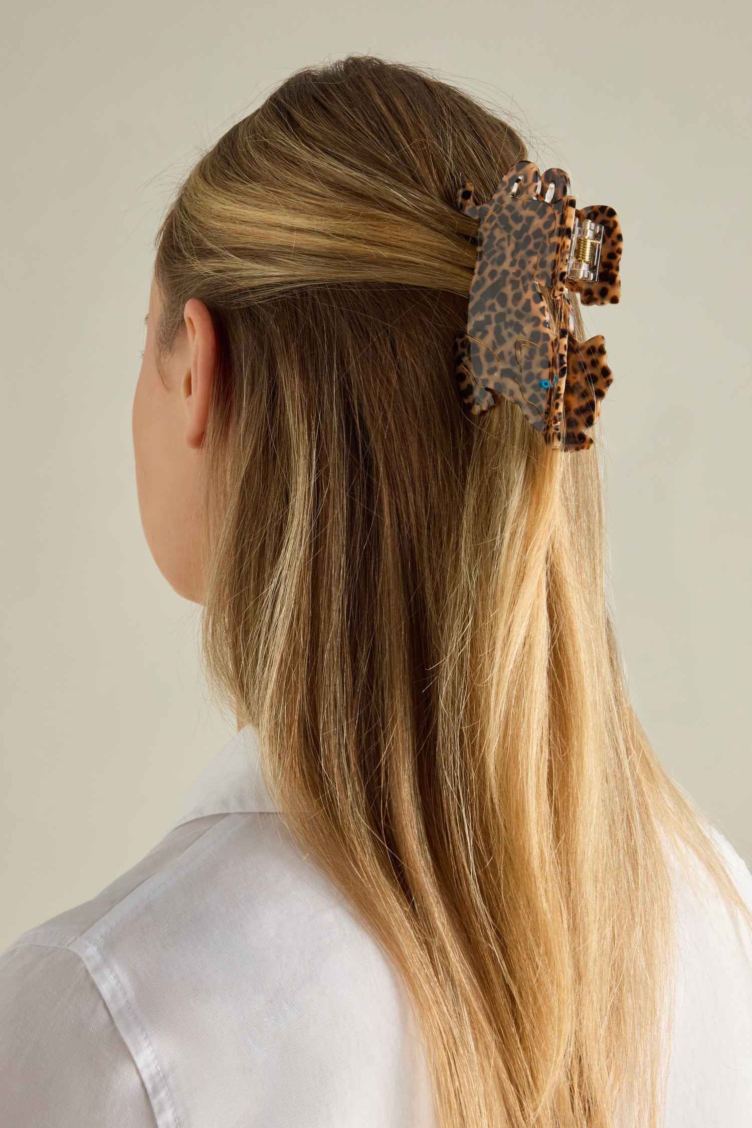 A woman with long blonde hair, adorned with a Hand Painted Leopard Hair Clip crafted from biodegradable acetate, wears a white shirt and is facing away.