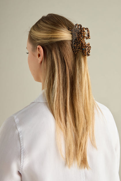 A woman with long blonde hair, wearing a white shirt, is viewed from the back. Her hair is partially secured with a Hand Painted Leopard Hair Clip.