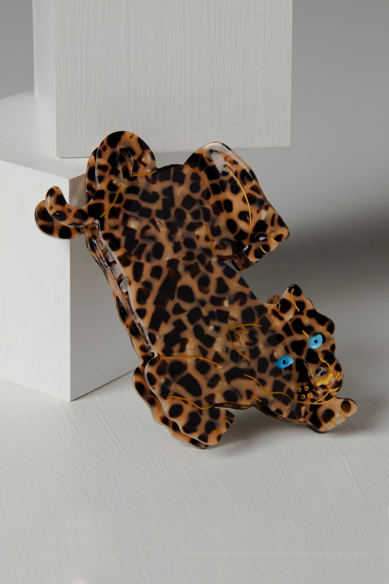 The Hand Painted Leopard Hair Clip, featuring blue eyes, rests against a white angular structure and showcases eco-friendly craftsmanship with biodegradable acetate.