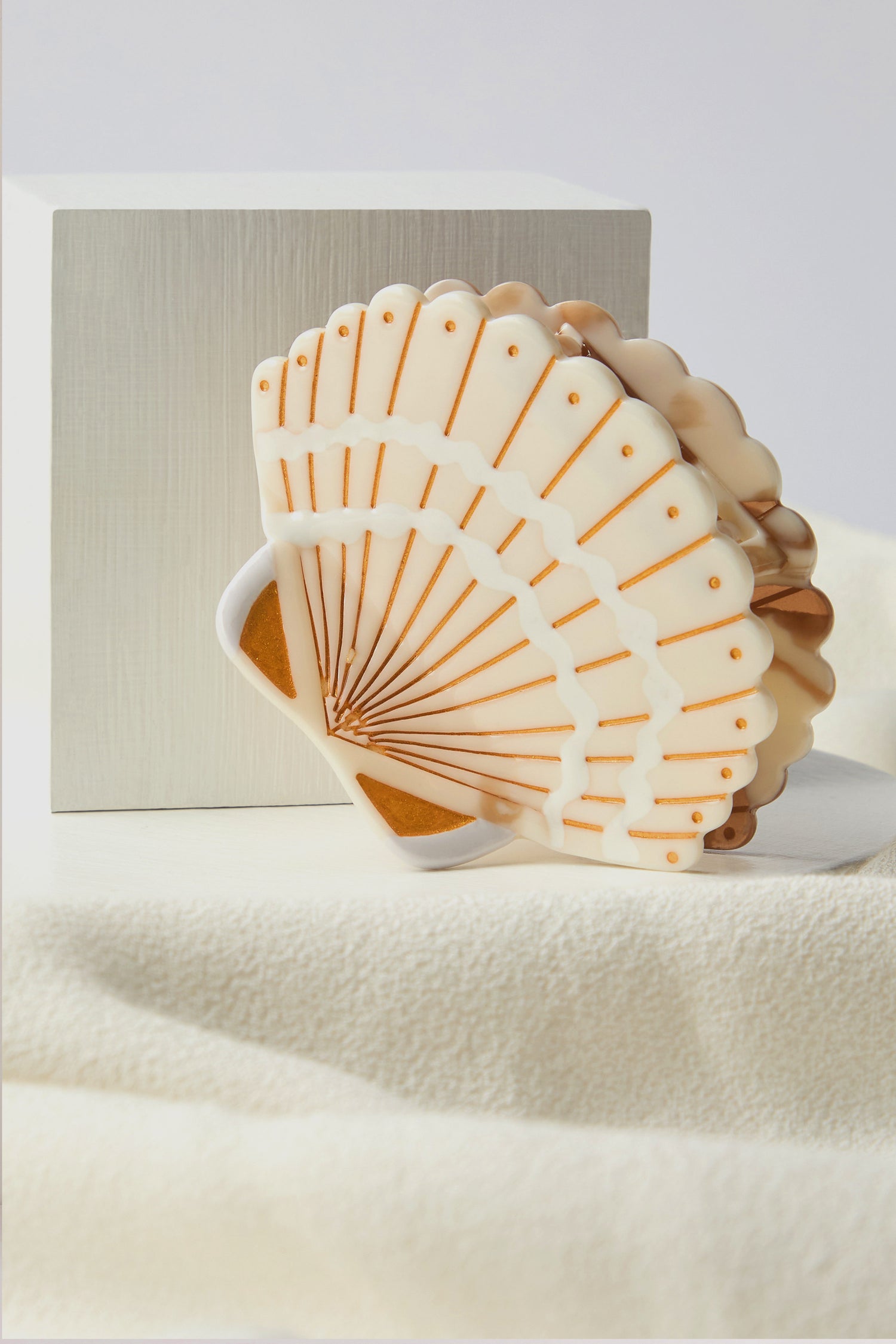 The Hand Painted Seashell Hair Clip, highlighting sustainable craftsmanship, is elegantly displayed against a light-colored cube and fabric surface.