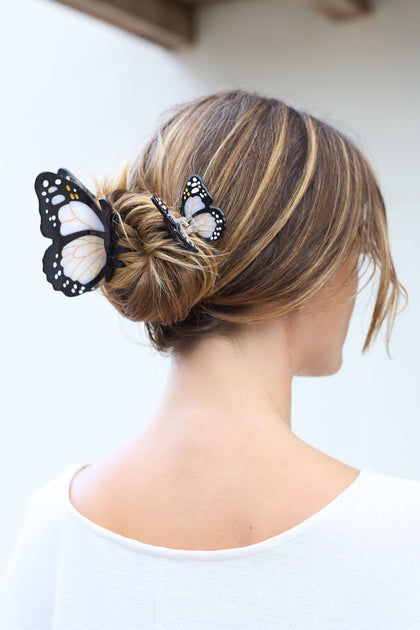 A woman with light brown hair in a bun wears two large hand-painted Mini Monarch Hair Clips, each featuring a beautiful monarch butterfly design. She faces away in a white top, accessorized with clips made from biodegradable acetate.