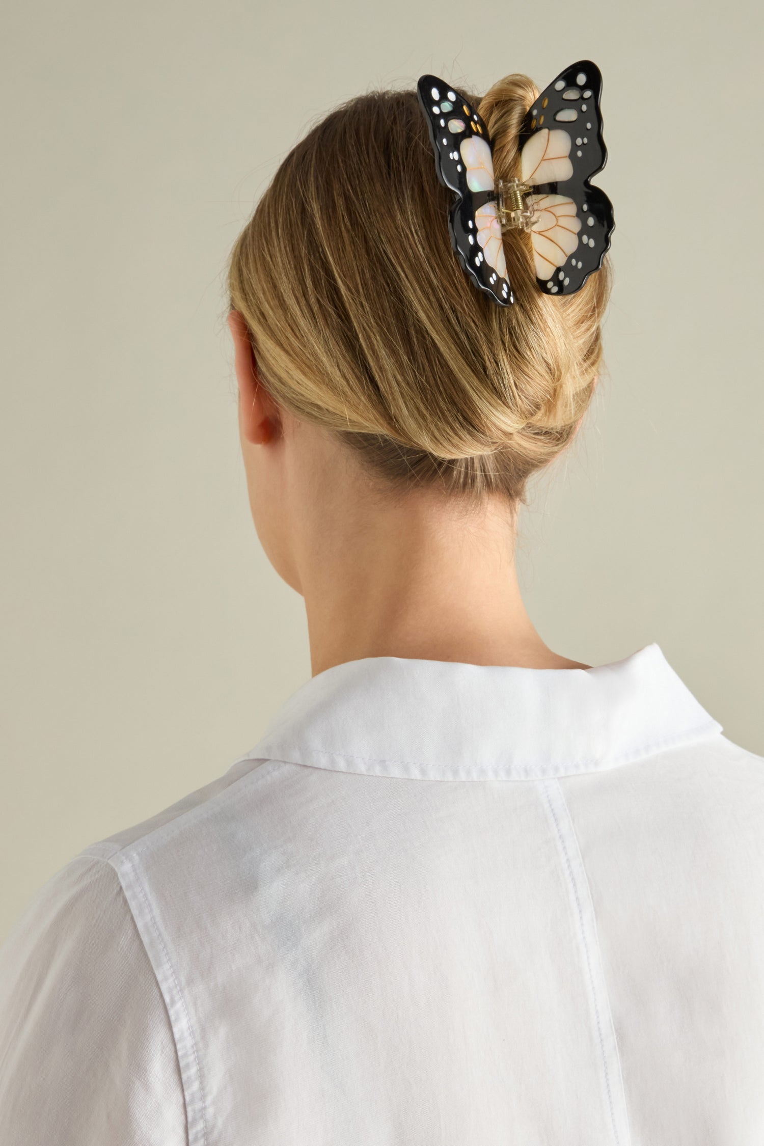 A woman with blonde hair is seen from behind, wearing a white collared shirt and securing her bun with a Hand Painted Monarch Butterfly Hair Clip made of biodegradable acetate.