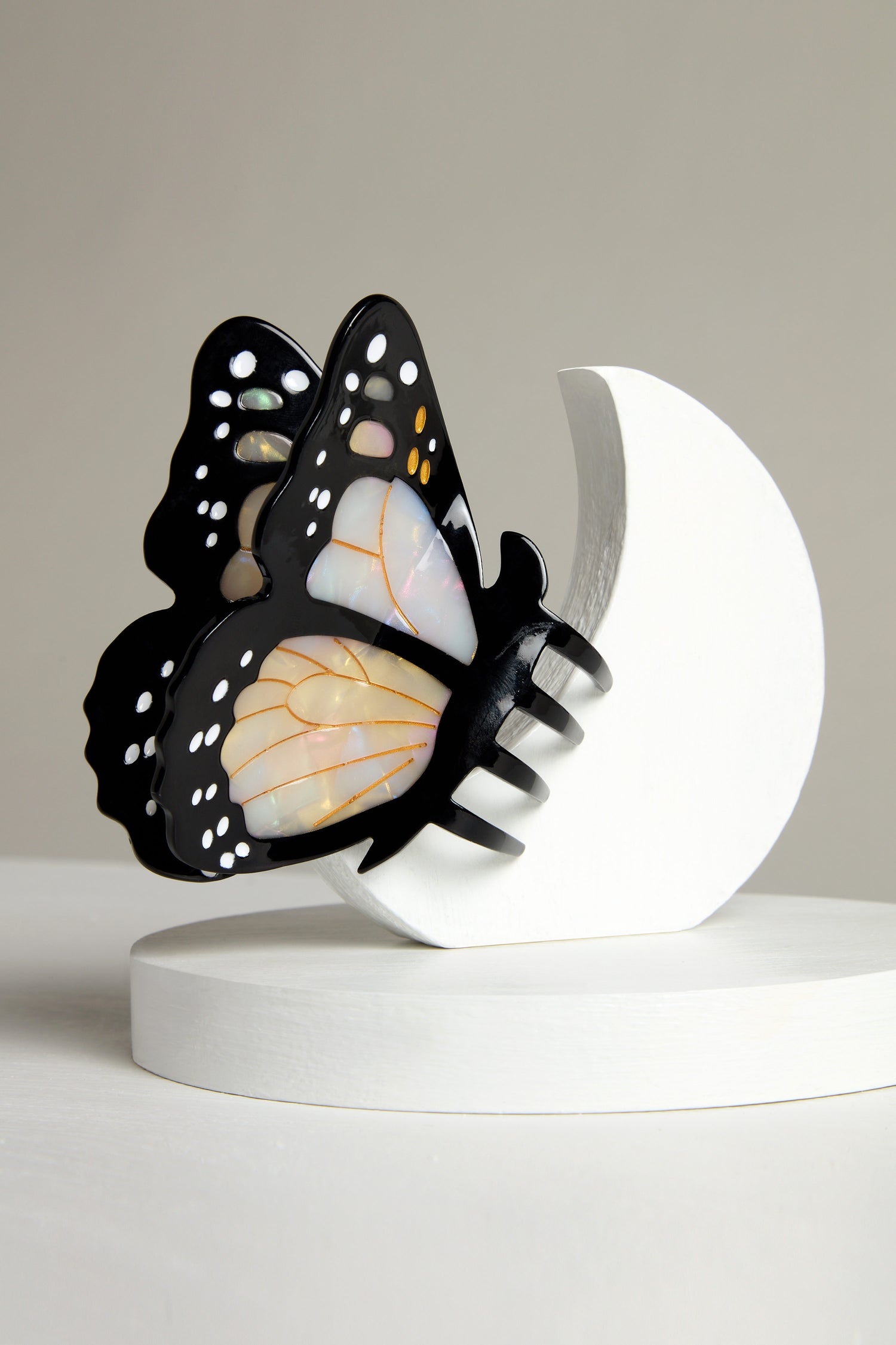 The Hand Painted Monarch Butterfly Hair Clip, crafted from biodegradable acetate, showcases intricate black and white patterns. It rests elegantly on a curved white stand against a neutral background, highlighting eco-friendly artistry.