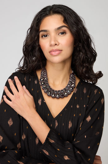 A woman with wavy dark hair wears a black patterned dress and the Velvet Ribbon Statement Necklace, posing with one arm across her chest against a plain background.