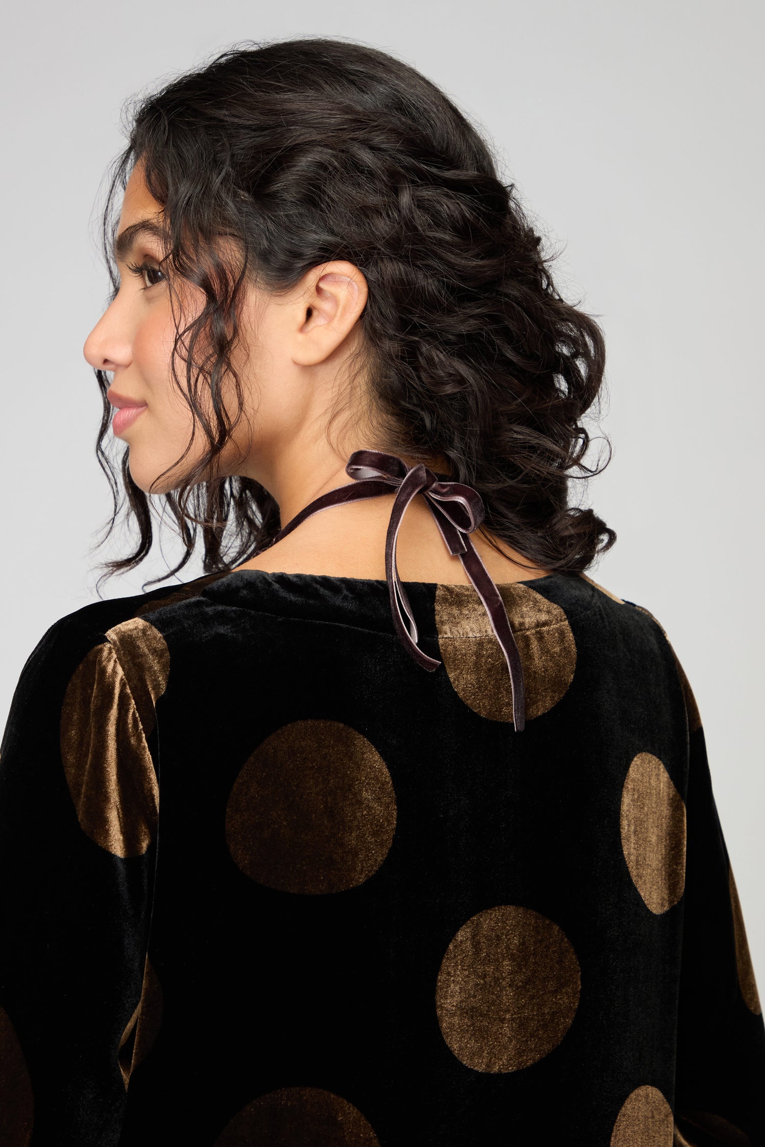 A person with dark curly hair wears the Velvet Ribbon Statement Necklace, paired with a black velvet top adorned with large brown polka dots and a tied ribbon at the back, creating a chic and stylish look.