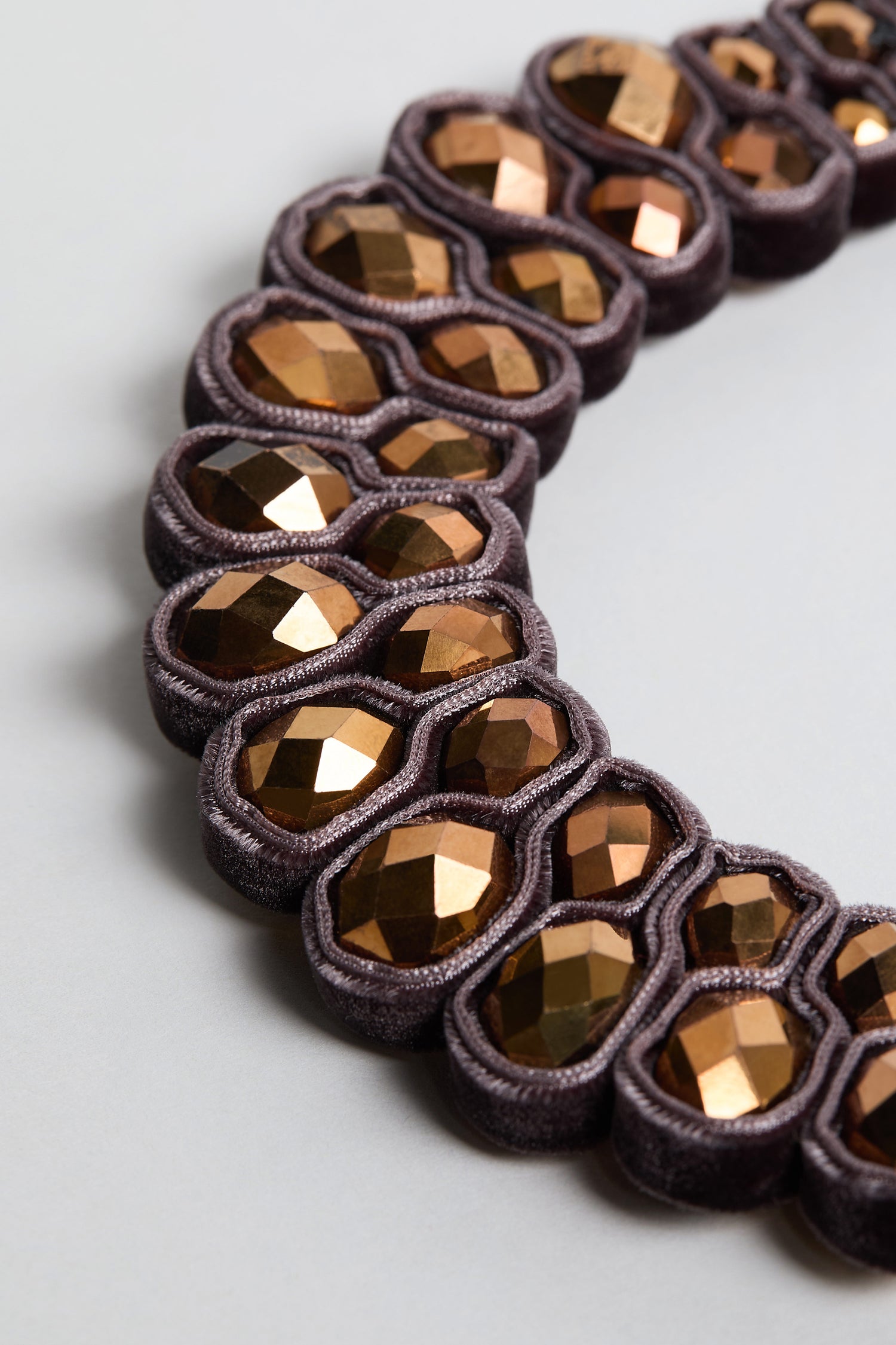 A close-up of the Velvet Ribbon Statement Necklace, showcasing faceted metallic bronze beads set in dark purple velvet loops, arranged in a curved pattern on a light gray background.