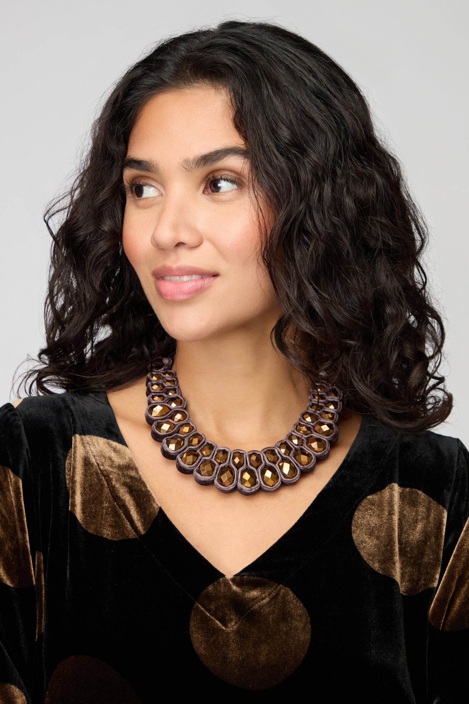 A woman with wavy dark hair wears a black velvet top with large gold polka dots and the Velvet Ribbon Statement Necklace, which features multiple gold and brown elements. She looks slightly to the side.