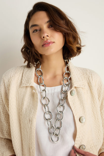 A woman with short brown hair wears a textured cream jacket, white top, and the Long Links Necklace, standing against a plain light background.