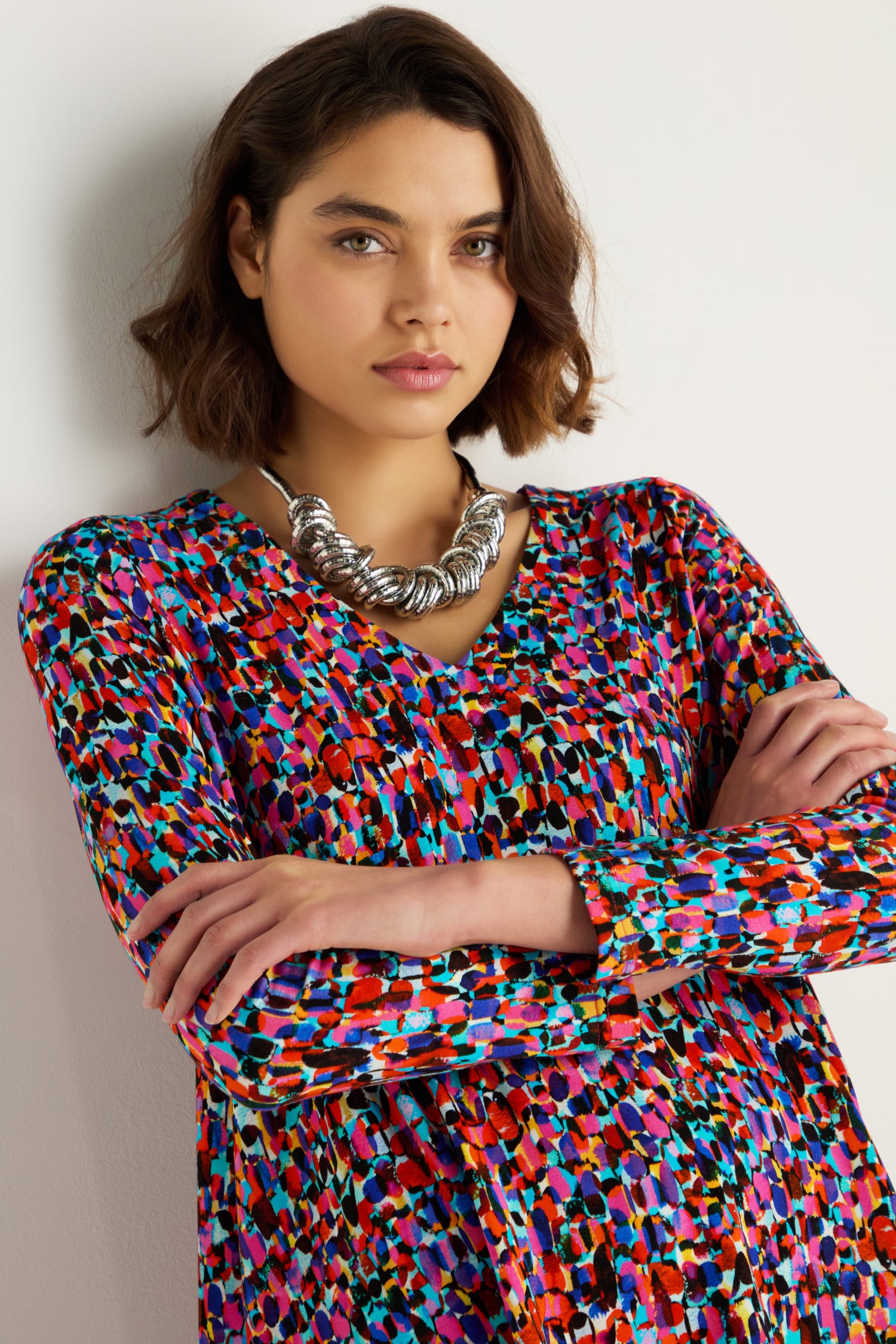 A woman with short brown hair, wearing a colorful patterned top and the Woven Silver Necklace, stands with arms crossed against a plain light background.