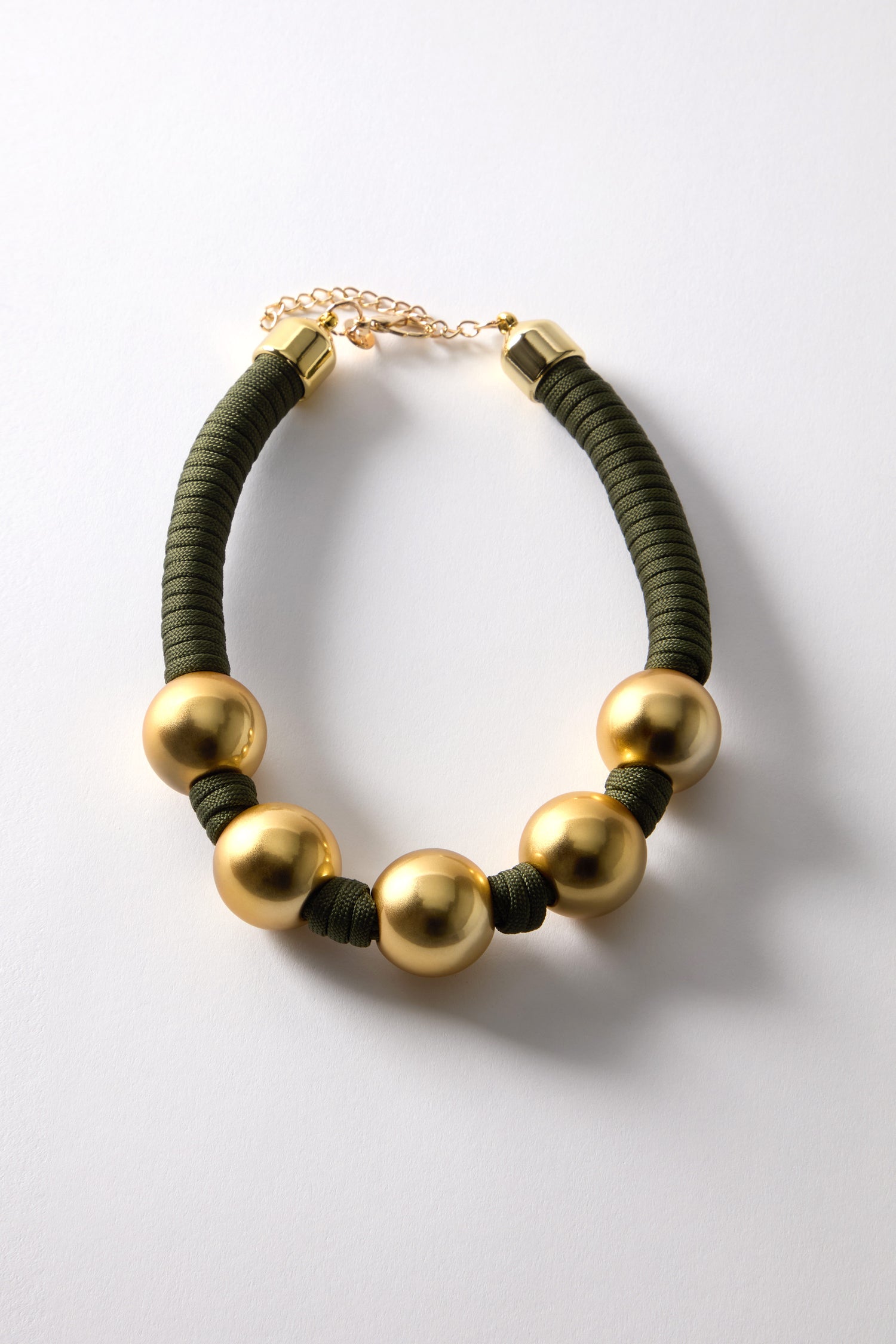 The Gold Sphere Cord Necklace features large gold spheres on a khaki green braided cord, paired with a gold chain clasp against a white backdrop.