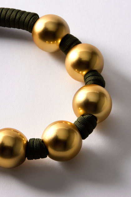 The Gold Sphere Cord Necklace displays smooth, shiny gold spheres on a khaki green rope, arranged in a partial circle against a white background.