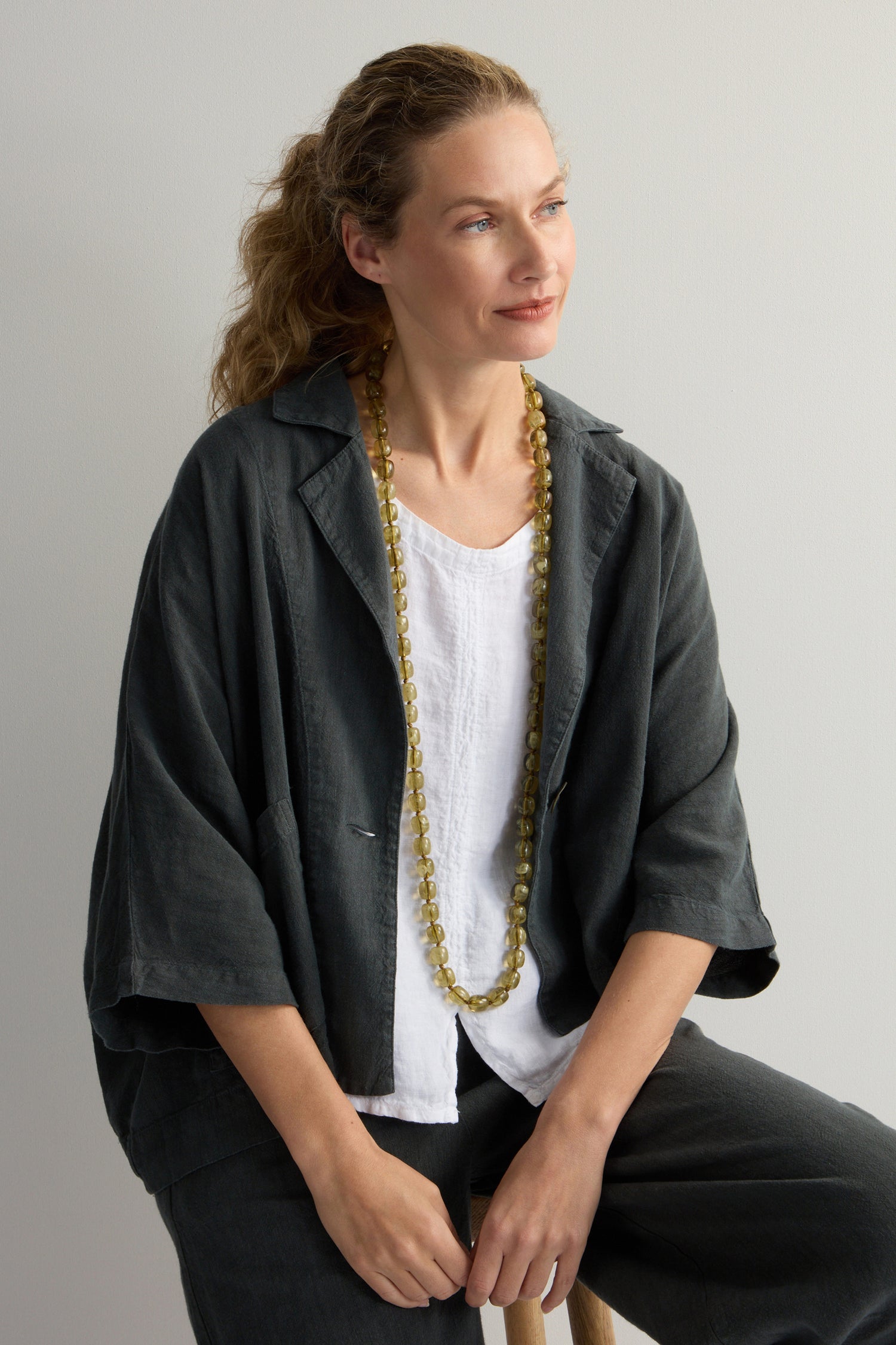 A woman with wavy hair sits on a stool, wearing a dark jacket over a white top and accessorized with an earthy khaki Long Resin Bead Necklace. She gazes to the side with a neutral expression against a plain background.