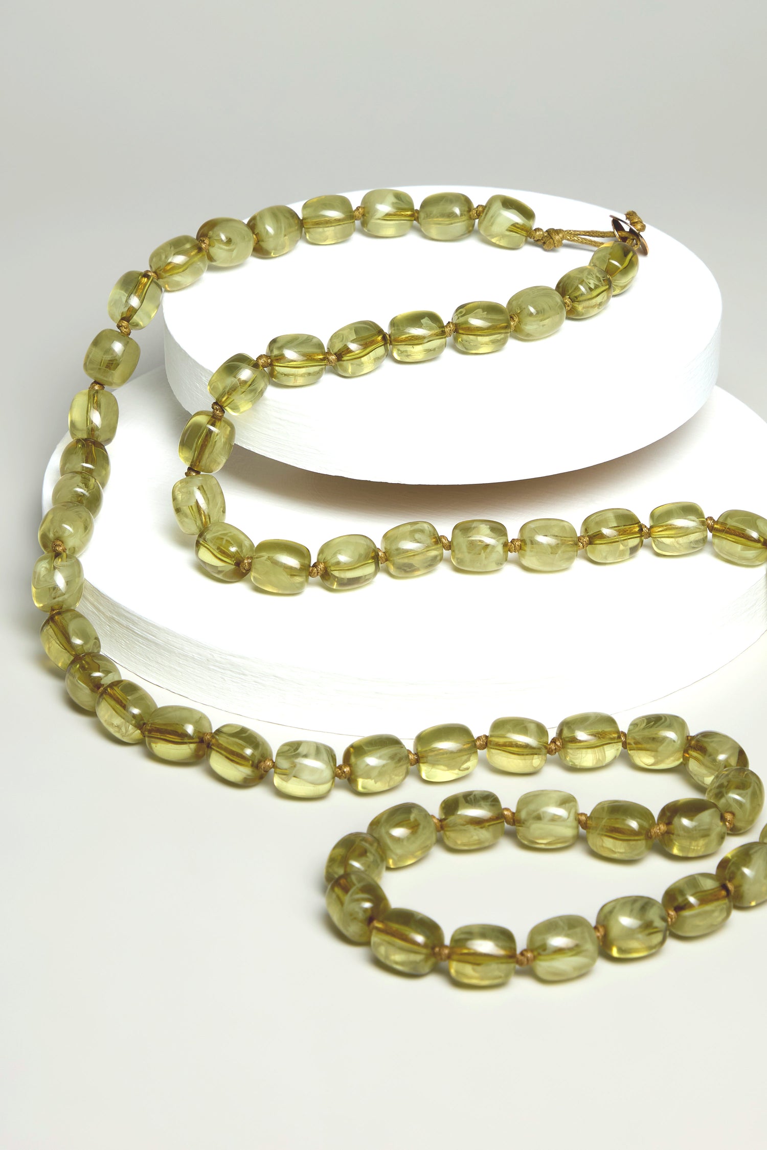 The Long Resin Bead Necklace, featuring translucent green beads, is artistically displayed on round white platforms. Its earthy khaki tones make it a versatile accessory that complements any style.