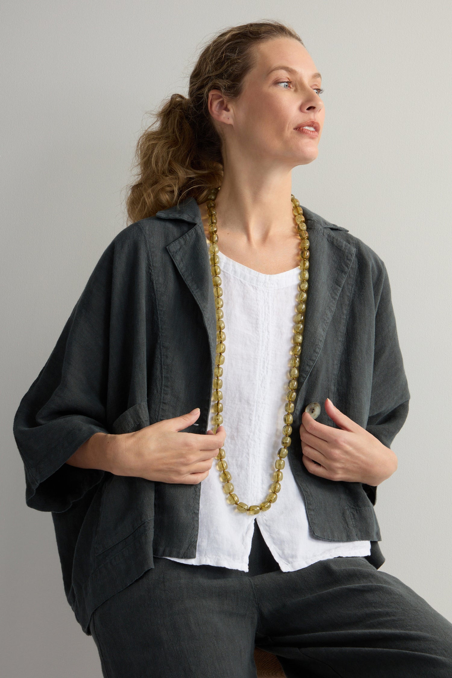 A woman in a gray jacket over a white top wears the Long Resin Bead Necklace in earthy khaki tones while looking to the side against a plain background. This versatile piece adds subtle elegance to her ensemble.