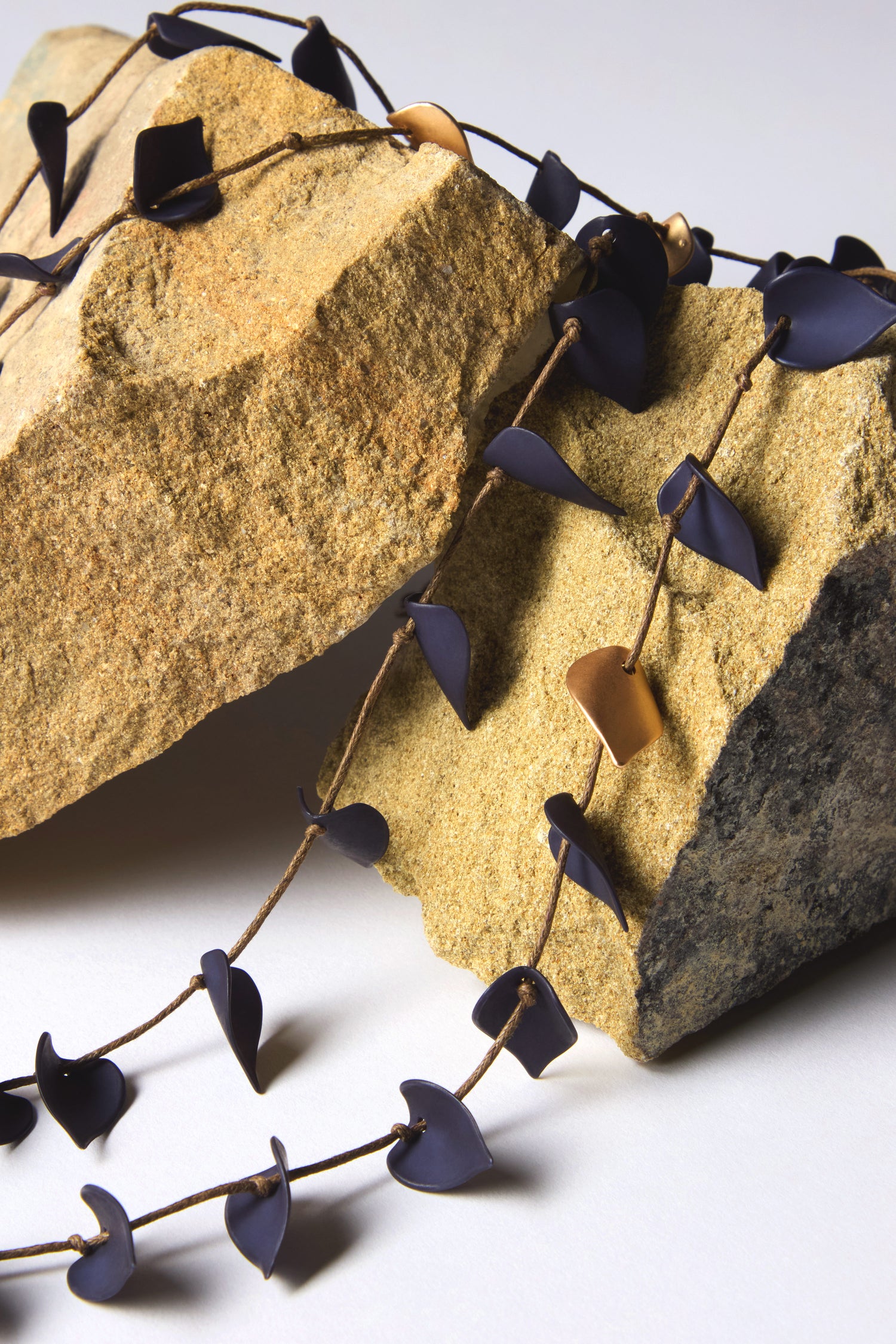 A Navy Cascade Leaves Necklace with dark and gold-toned leaf-shaped elements elegantly rests on two large, rough stones.