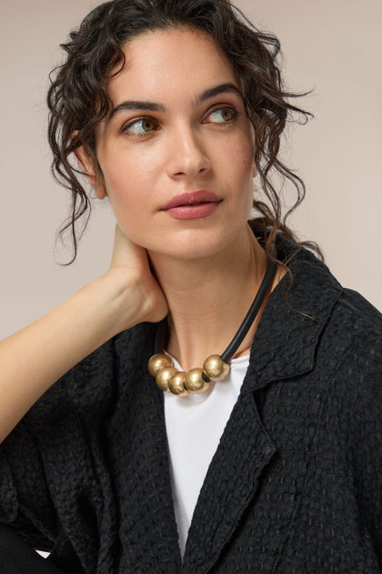 A woman wearing a black jacket and Hammered Gold Spheres Necklace with black cord.