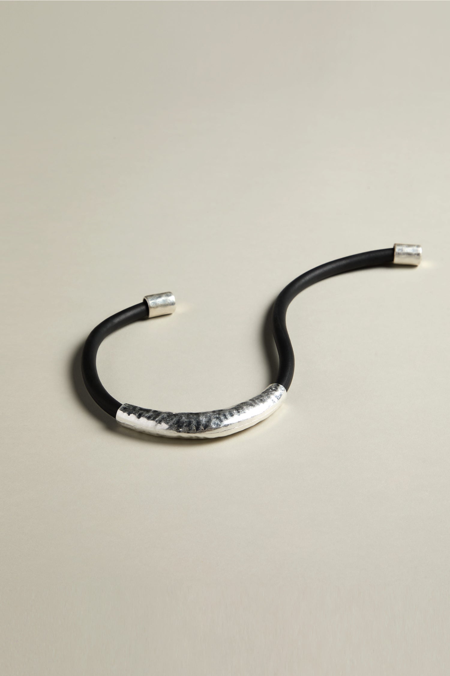 A Hammered Necklace, featuring a black and silver open bracelet with a textured silver section that resembles a hammered tubular pendant, is lying on a light surface.