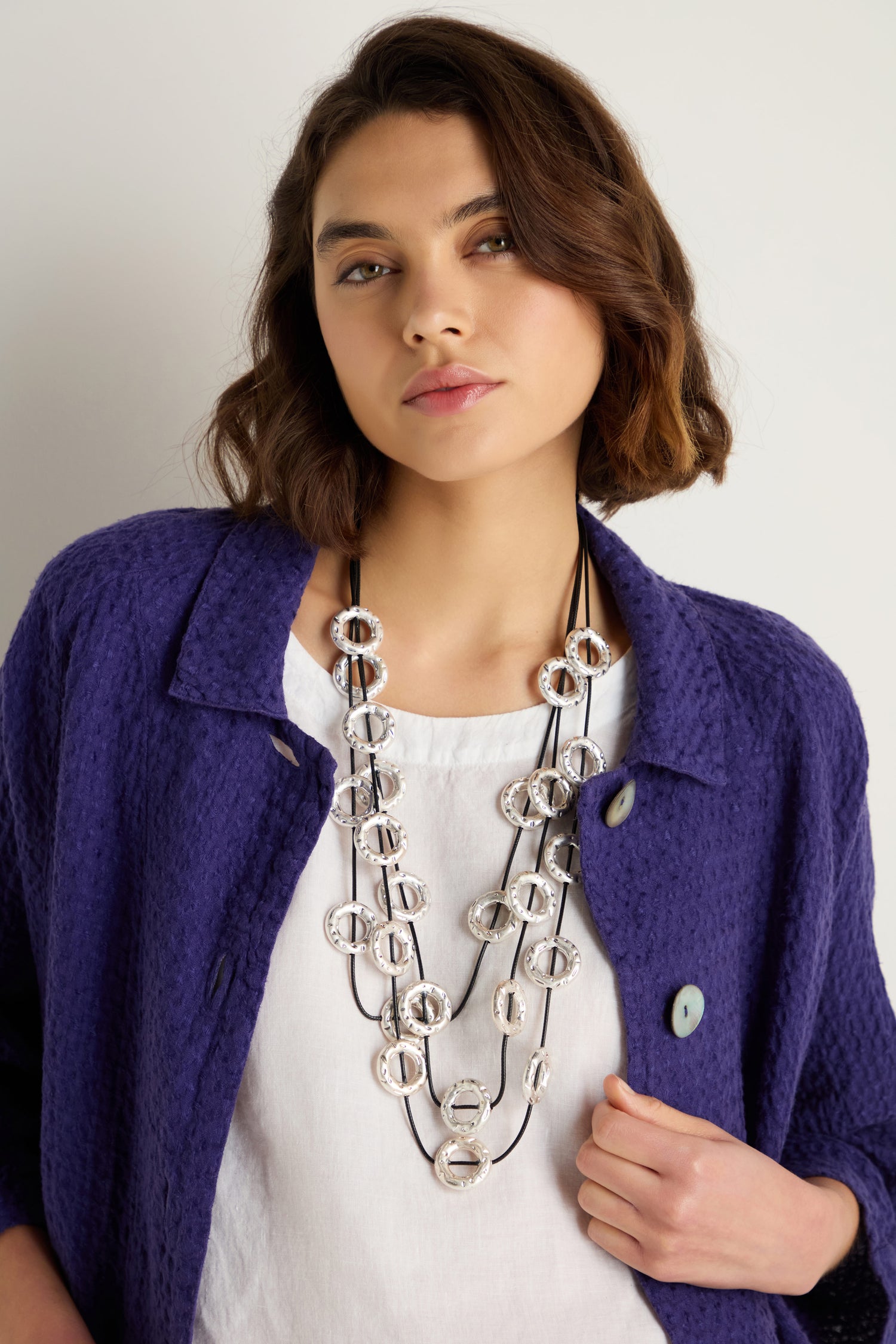 A woman with short brown hair wears a textured purple jacket, white top, and the Cascade Hoops Necklace, standing against a plain light background.