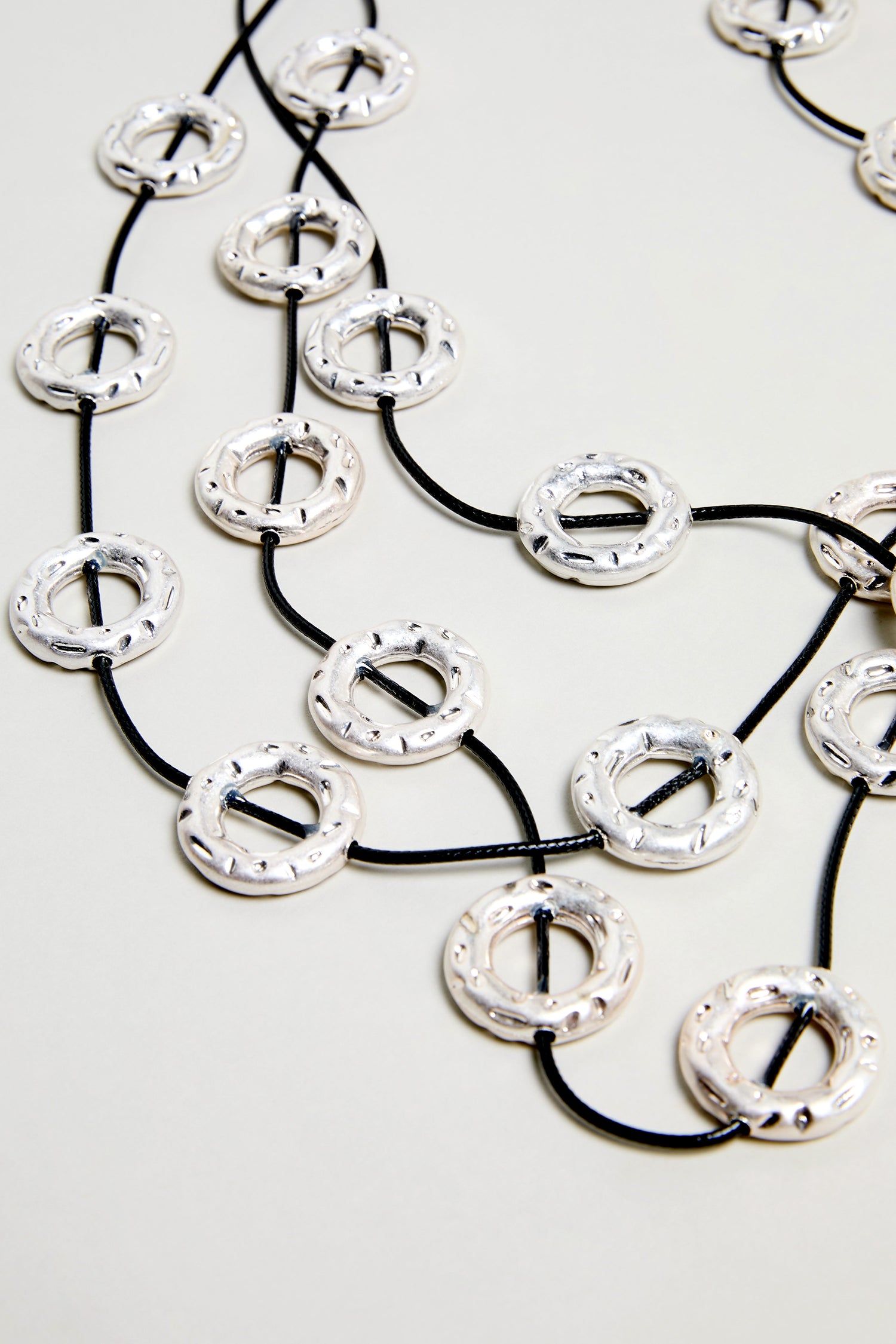 The Cascade Hoops Necklace showcases layered silver hoops with a textured finish, joined by black cords and arranged in overlapping circles against a light background.