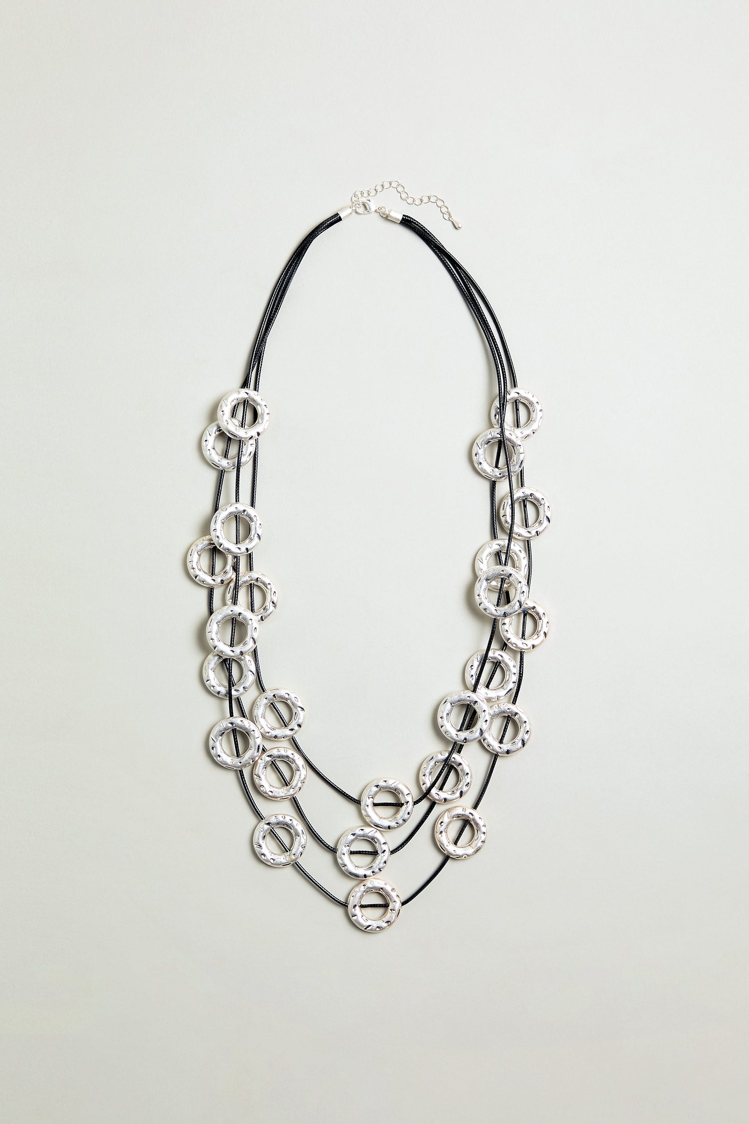 The Cascade Hoops Necklace features three black cords and layered silver textured metal hoops, shown against a plain light background.