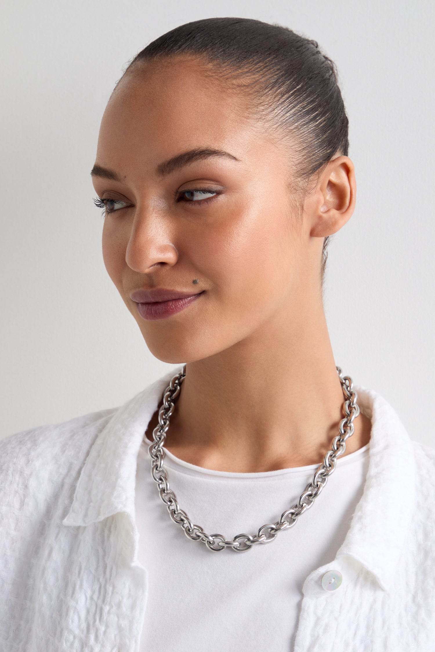 A person with their hair tied back and a white shirt wears a Short Silver Chain Necklace from the versatile jewellery collection, looking to the side against a plain background.