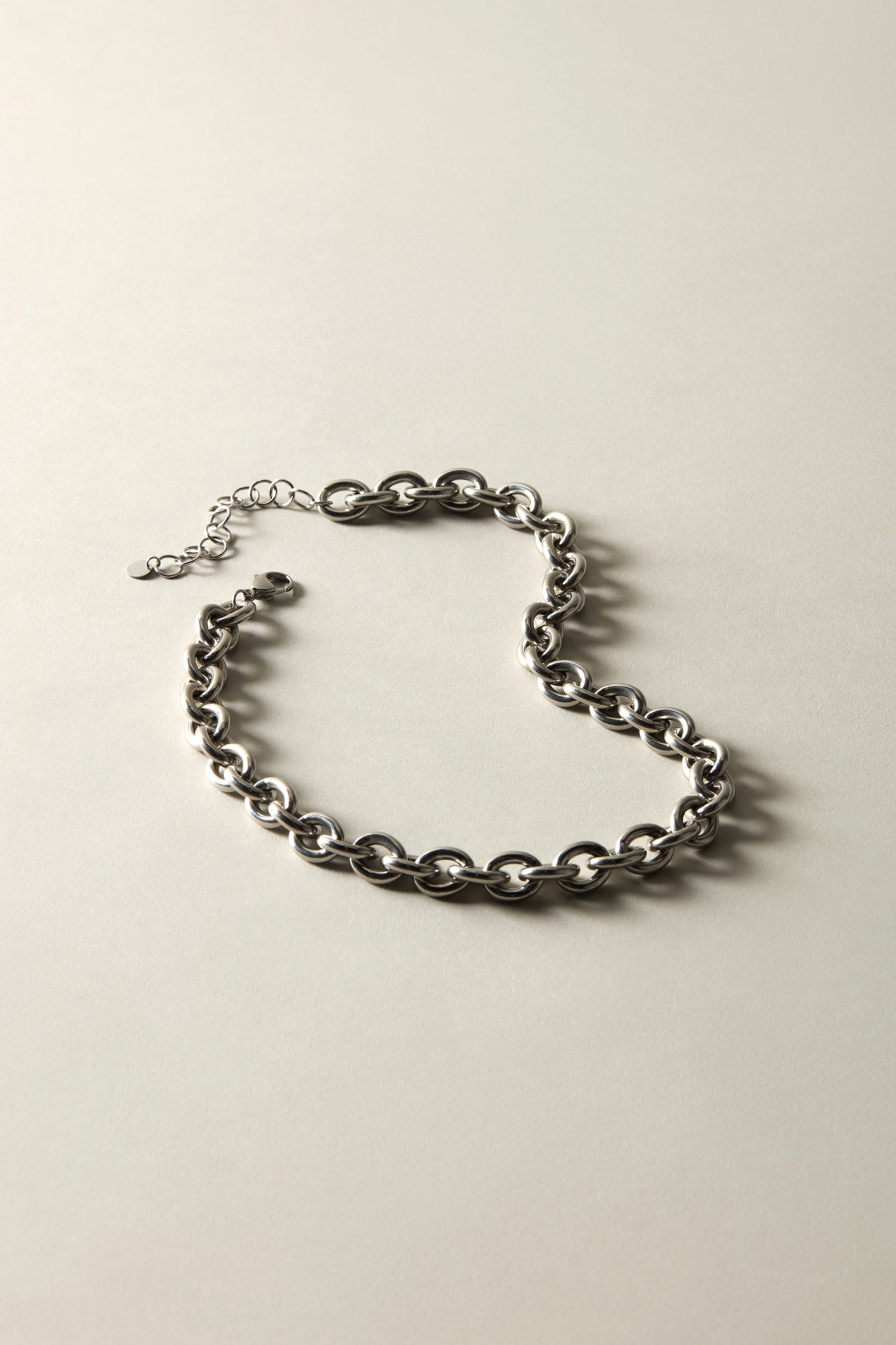 The Short Silver Chain Necklace, featuring large round links and a versatile design, is elegantly arranged in an S shape on a light gray surface—an ideal addition to elevate your jewelry collection.