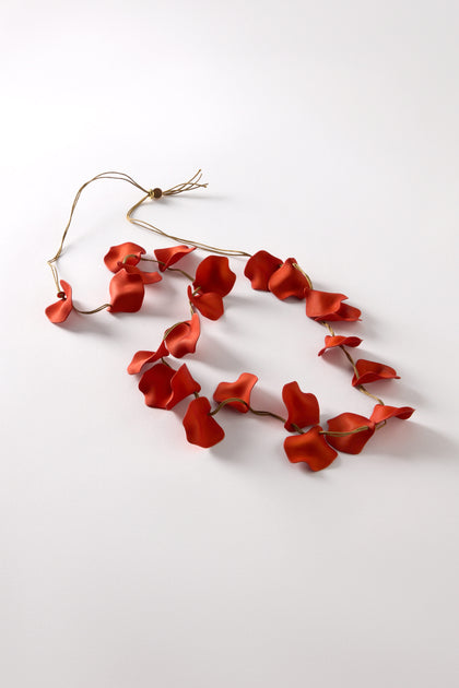 A Long Cascade Petal Necklace, composed of red petal-shaped pieces linked by a thin flexible wire, is elegantly displayed on a white surface. Its coral color adds vibrancy, and the adjustable cord ensures a perfect fit.