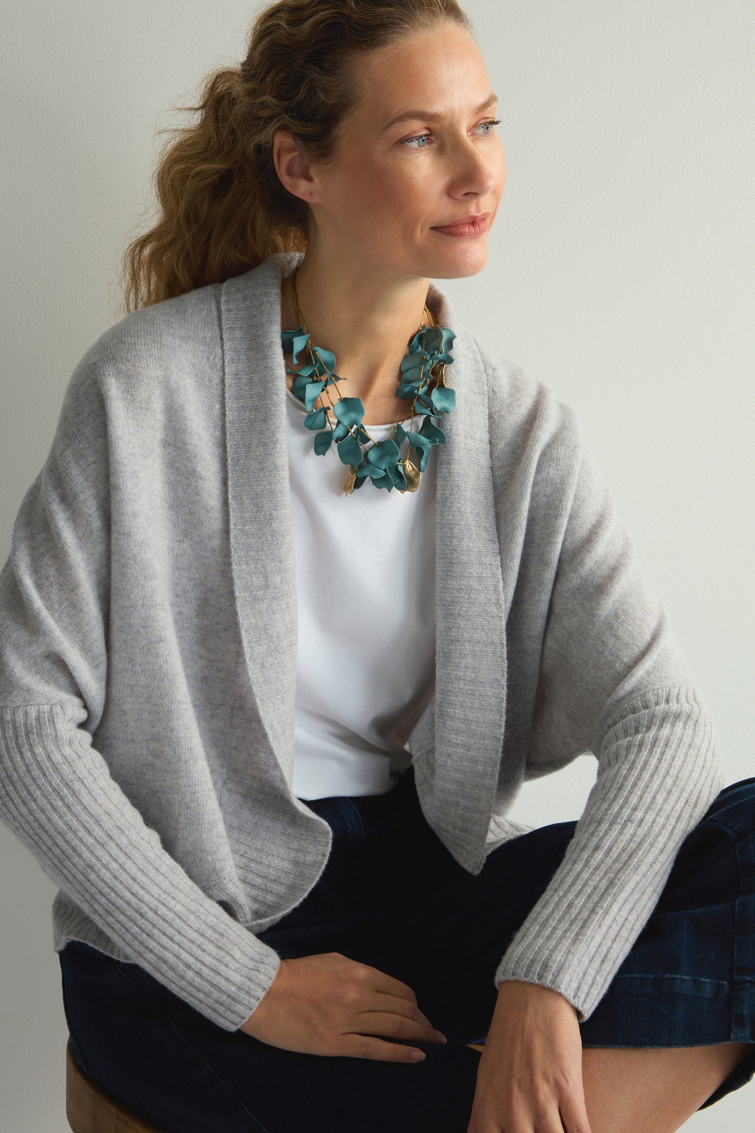 A person in a light gray cardigan, adorned with the Short Cascade Petal Necklace, sits gazing to the side against a plain backdrop. The necklace's organic elegance subtly enhances their appearance with charming sophistication.