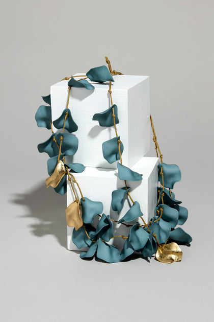 The Short Cascade Petal Necklace, with teal and gold leaf-shaped elements, elegantly drapes over two white cube pedestals. Its adjustable cord complements the sculptural beauty against a neutral backdrop, showcasing organic elegance.