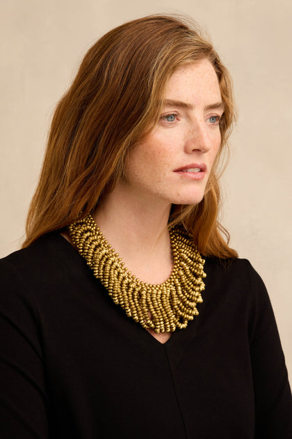 A woman with wavy auburn hair, dressed in a black top, wears the Gold Beaded Collar Necklace—a large, multi-strand accessory—and looks slightly to the right against a neutral background.