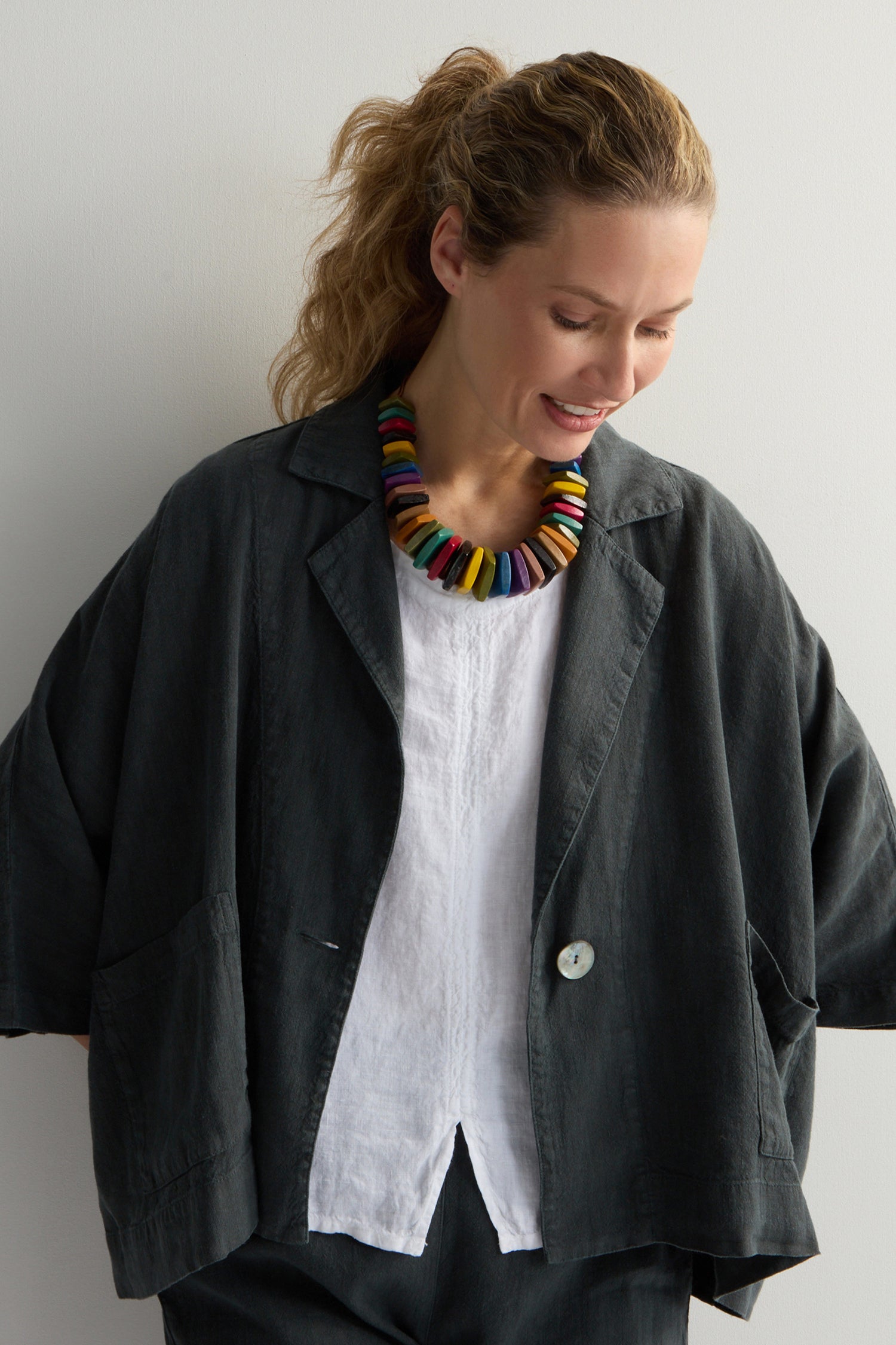 A person in a dark oversized jacket and white shirt stands against a plain backdrop, sporting the Rainbow Wooden Bead Cord Necklace, hands casually tucked into pockets.