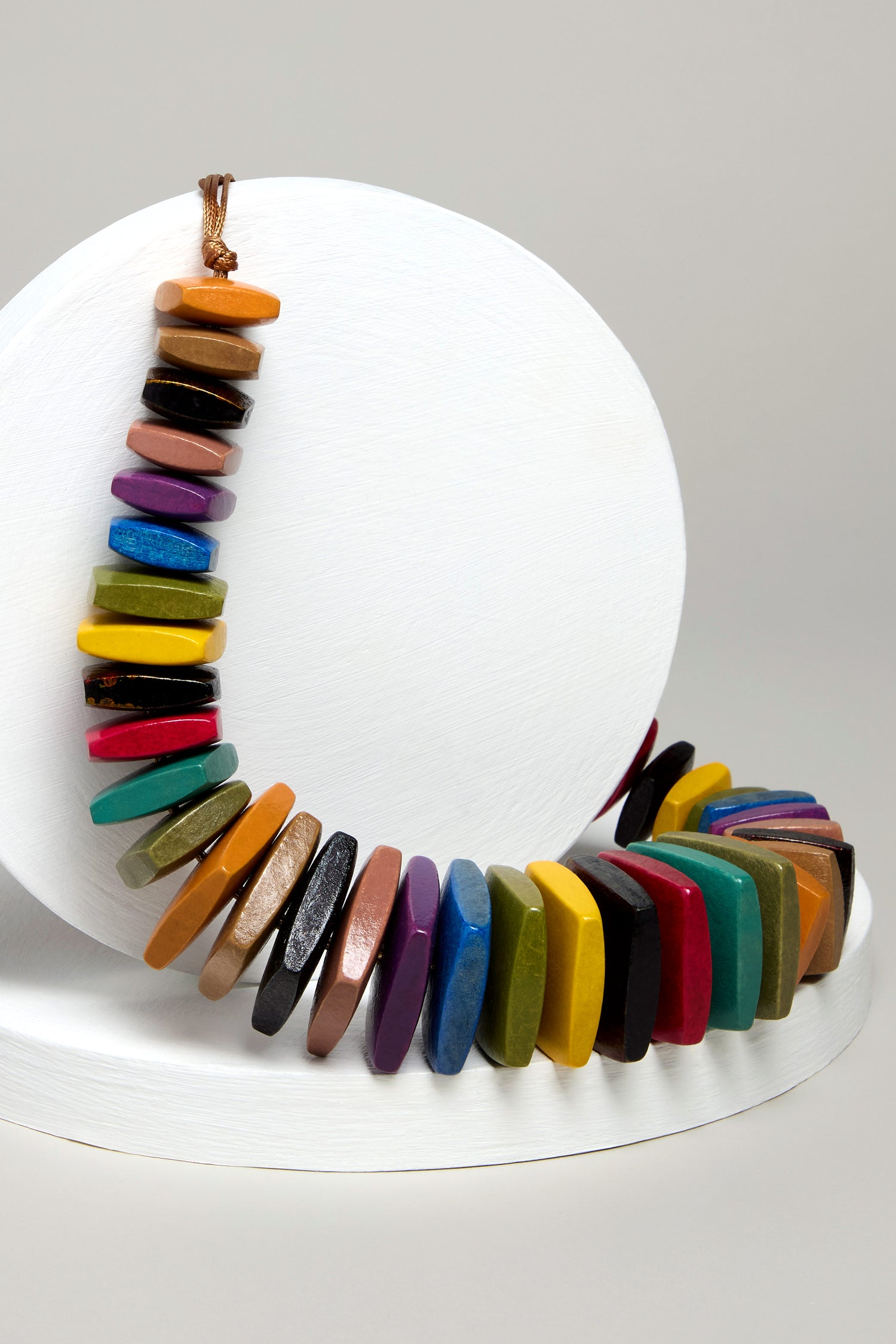 The Rainbow Wooden Bead Cord Necklace, with flat oval beads in assorted colors, is elegantly showcased on a white round stand set against a gray background.