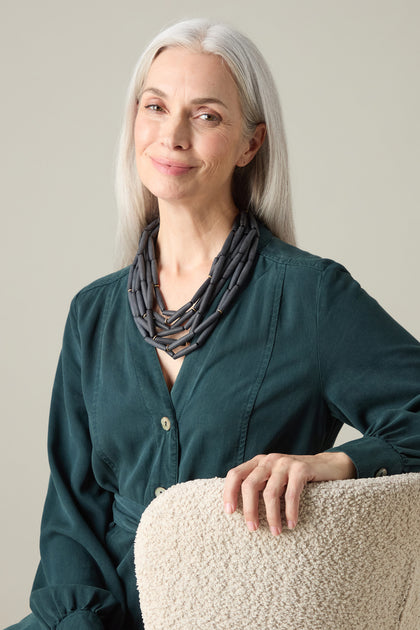 An older woman with long gray hair, dressed in a dark green blouse and wearing the Noir Cascade Necklace with matte black beads, is seated on a textured chair, smiling softly.