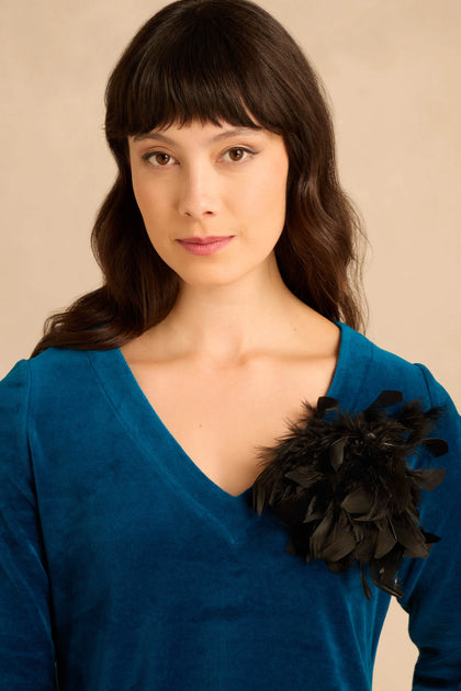 A woman wears a blue V-neck top accessorized with a black Feather Corsage pinned to her chest.