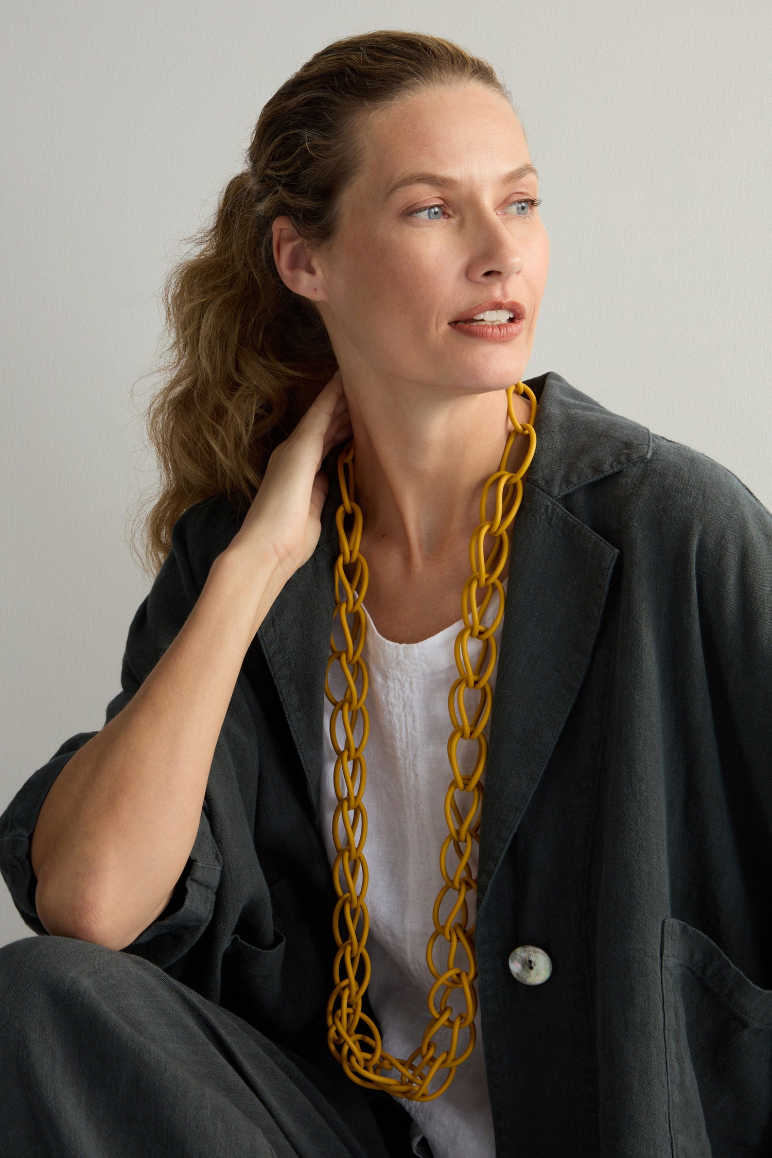 A woman in a dark oversized blazer and white top wears the Mustard Long Links Necklace, a modern and lightweight accessory, while looking to the side.