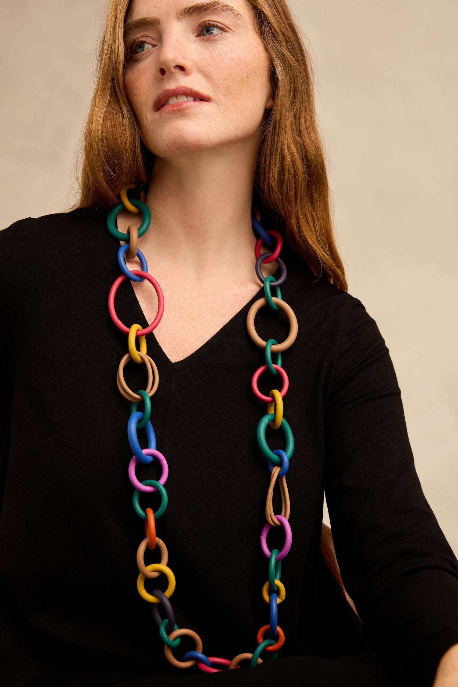 A woman with long brown hair wears a black top and a Long Rainbow Hoops Necklace, which features large, colorful interlocking rubber links for a striking effect.