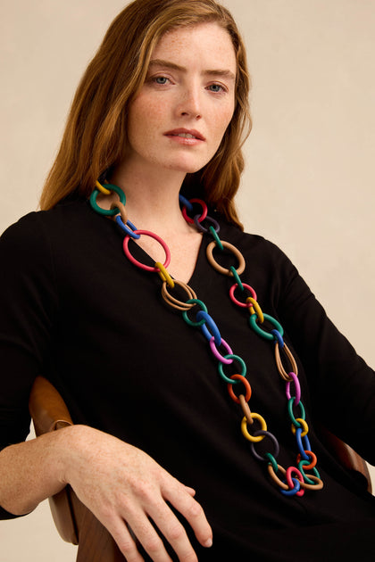 A woman wearing a black top sits on a chair, showcasing the Long Rainbow Hoops Necklace—an eye-catching accessory featuring oversized rubber links in vibrant colors—against a neutral background.