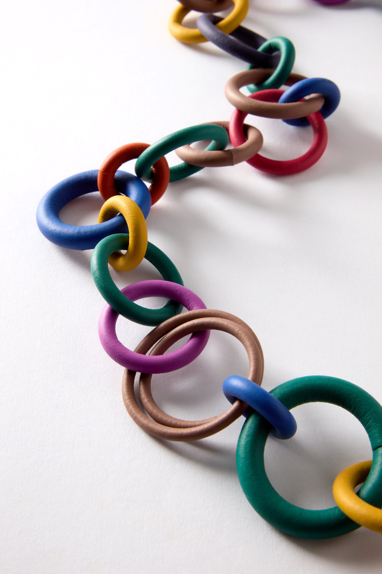 The Long Rainbow Hoops Necklace features a lively chain of interlinked hoops in blue, red, yellow, green, and brown that dances playfully against a white background.