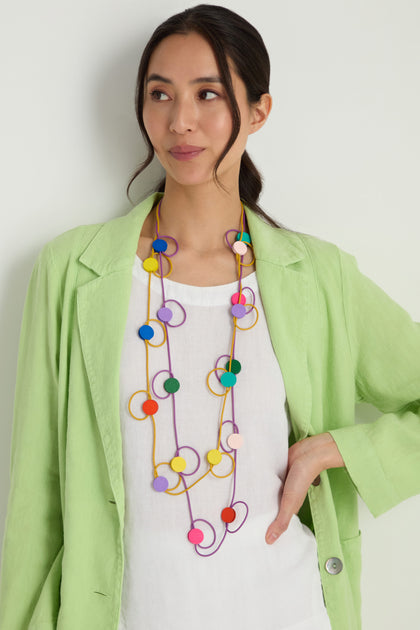 A woman in a light green blazer and white blouse wears the Rainbow Circle Hoop Necklace, featuring colorful circular beads, standing against a plain white background.