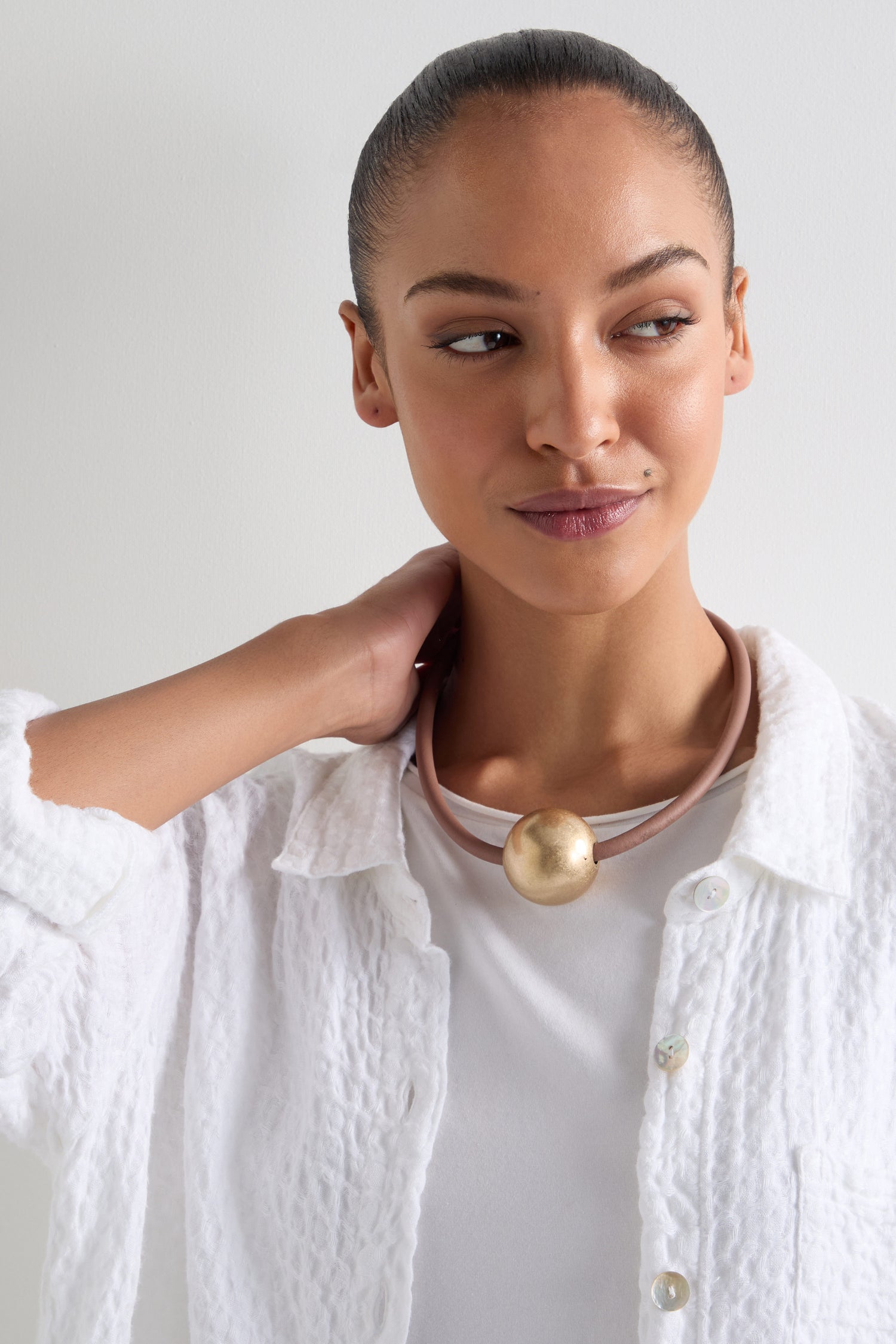 Wearing a textured white shirt, the individual gazes sideways with their hand on their neck, showcasing the Single Sphere Necklace. The necklace's minimalist design and large round pendant reflect contemporary style.