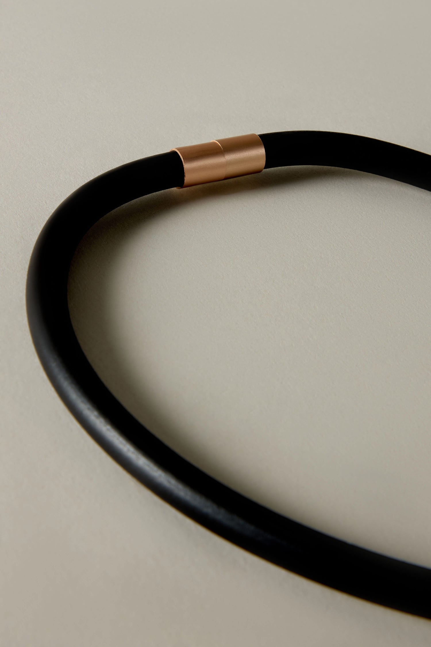 A close-up of the Single Sphere Necklace, featuring a sleek black circular band with a copper-hued connector, resting on a light gray surface, highlighting its contemporary design and minimalist appeal.